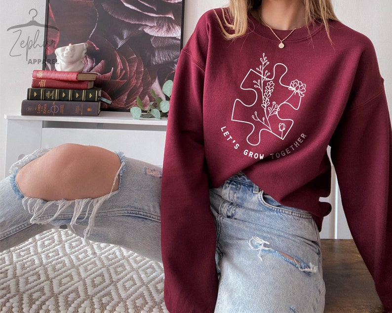May include: A maroon sweatshirt with a white outline of a puzzle piece with flowers inside. The text "Let's Grow Together" is written around the puzzle piece.