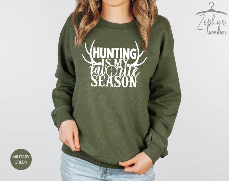 K&ouml;nnte beinhalten: Ein gr&uuml;ner Pullover mit dem Text "Hunting is my favorite season" in Wei&szlig; mit einem wei&szlig;en Ziel und Geweih.
