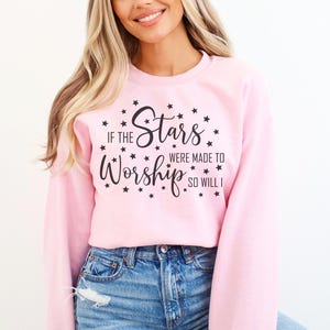 May include: A light pink sweatshirt with the text "If the Stars Were Made to Worship So Will I" in black script and star accents. The sweatshirt is paired with blue denim jeans.