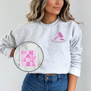 May include: A gray sweatshirt with the text "In My Law School Era" in pink letters on the left side and the text "Boston University" with a pink gavel on the right side.