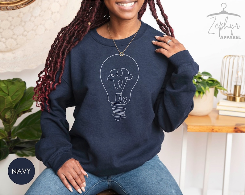 May include: A navy blue sweatshirt with a white outline of a lightbulb with a puzzle piece inside. The text "Zephyr Apparel" is in the top right corner of the image.