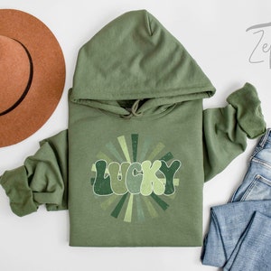 Peut inclure: Un sweat-shirt &agrave; capuche vert avec un motif r&eacute;tro du mot "LUCKY" dans un design en forme de soleil. Le sweat-shirt est pos&eacute; sur une surface blanche avec un chapeau marron et un jean bleu.