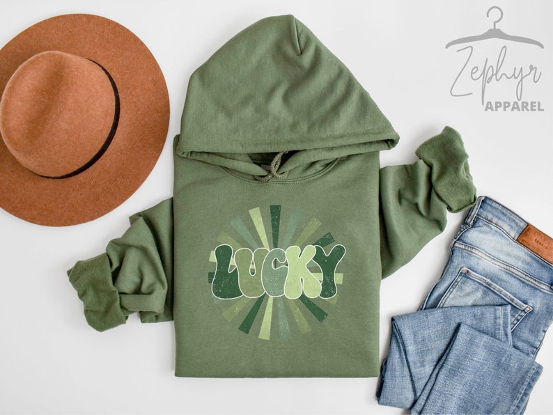 Peut inclure: Un sweat-shirt &agrave; capuche vert avec un motif r&eacute;tro du mot "LUCKY" dans un design en forme de soleil. Le sweat-shirt est pos&eacute; sur une surface blanche avec un chapeau marron et un jean bleu.