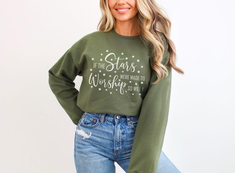 May include: Olive green sweatshirt with the text "If the Stars Were Made to Worship, So Will I" in white. The design includes star accents. The long-sleeved top is paired with blue jeans.