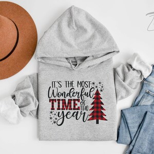 Puede incluir: Sudadera gris con capucha con el texto "It's the most Wonderful Time of the year" y un gr&aacute;fico de &aacute;rbol de Navidad rojo y negro a cuadros.