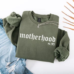 Op de afbeelding: Een groen sweatshirt met de tekst "motherhood est. 2025" in witte gotische letters.