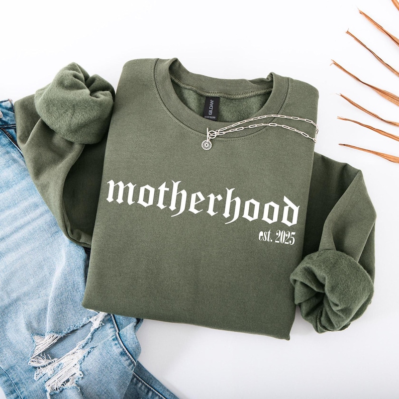 Op de afbeelding: Een groen sweatshirt met de tekst "motherhood est. 2025" in witte gotische letters.