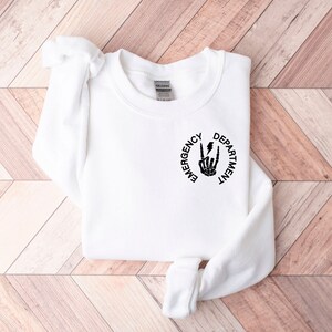 May include: White sweatshirt with a black graphic print of a skeleton hand making a rock and roll sign with a lightning bolt. The text "Emergency Department" is printed in a circle around the hand.