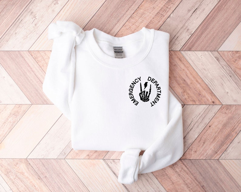 May include: White sweatshirt with a black graphic print of a skeleton hand making a rock and roll sign with a lightning bolt. The text "Emergency Department" is printed in a circle around the hand.