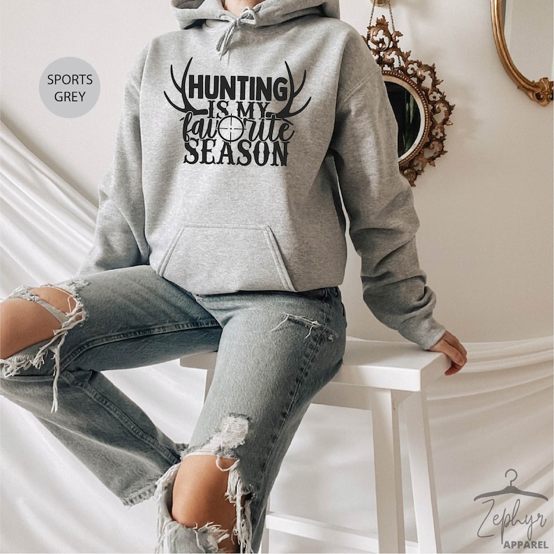 K&ouml;nnte beinhalten: Ein grauer Kapuzenpullover mit dem Text "Hunting is my favorite season" und Geweih auf beiden Seiten des Textes. Der Text ist in Schwarz und in der Mitte des Textes befindet sich ein Zielsymbol.
