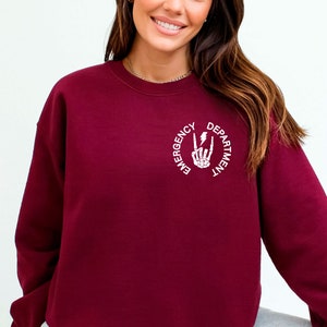 May include: Burgundy sweatshirt with a white graphic of a skeleton hand making a rock and roll sign with a lightning bolt. The text around the graphic reads "Emergency Department".