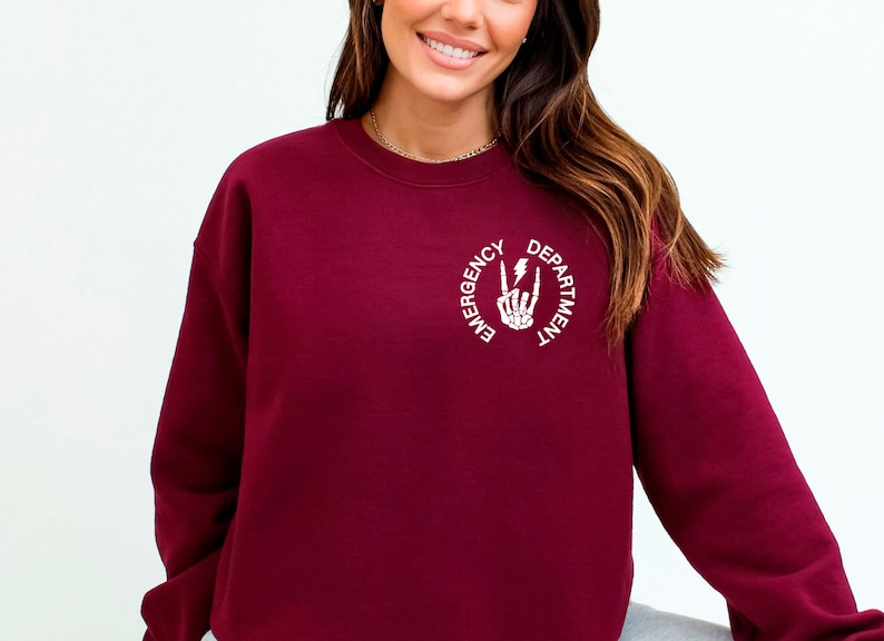 May include: Burgundy sweatshirt with a white graphic of a skeleton hand making a rock and roll sign with a lightning bolt. The text around the graphic reads "Emergency Department".