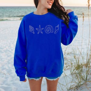 May include: A royal blue sweatshirt featuring a white outline design of seashells and a starfish. The sweatshirt is long-sleeved and has a crew neck. The background shows a beach scene with sand and ocean.