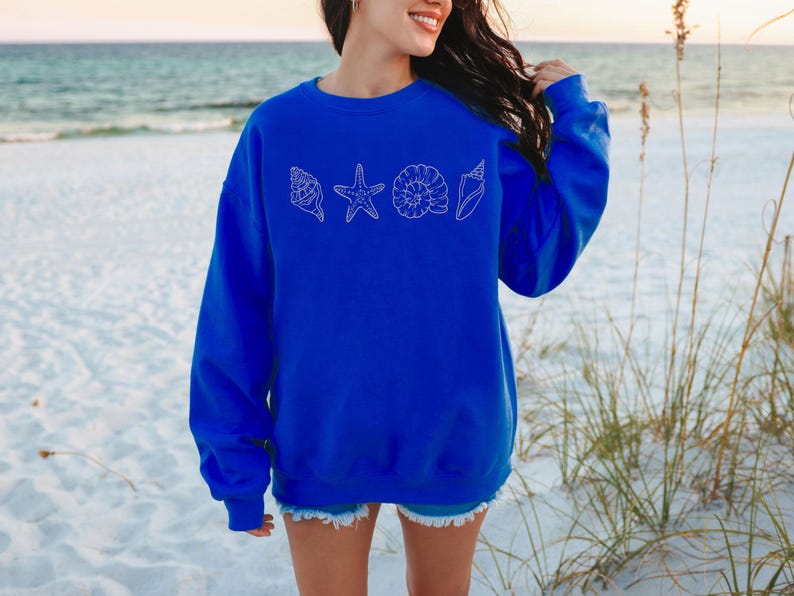 May include: A royal blue sweatshirt featuring a white outline design of seashells and a starfish. The sweatshirt is long-sleeved and has a crew neck. The background shows a beach scene with sand and ocean.