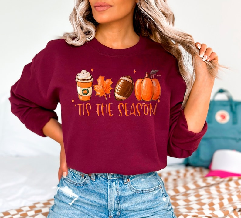 May include: Burgundy sweatshirt with a graphic of a coffee cup, a leaf, a football, and a pumpkin. The text "Tis the Season" is printed below the graphic.