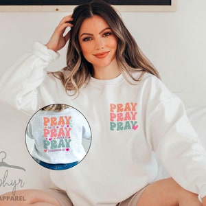 May include: White sweatshirt with a pink, orange and green graphic that says "Pray" three times. The graphic is on the front of the sweatshirt. The sweatshirt is being worn by a person.