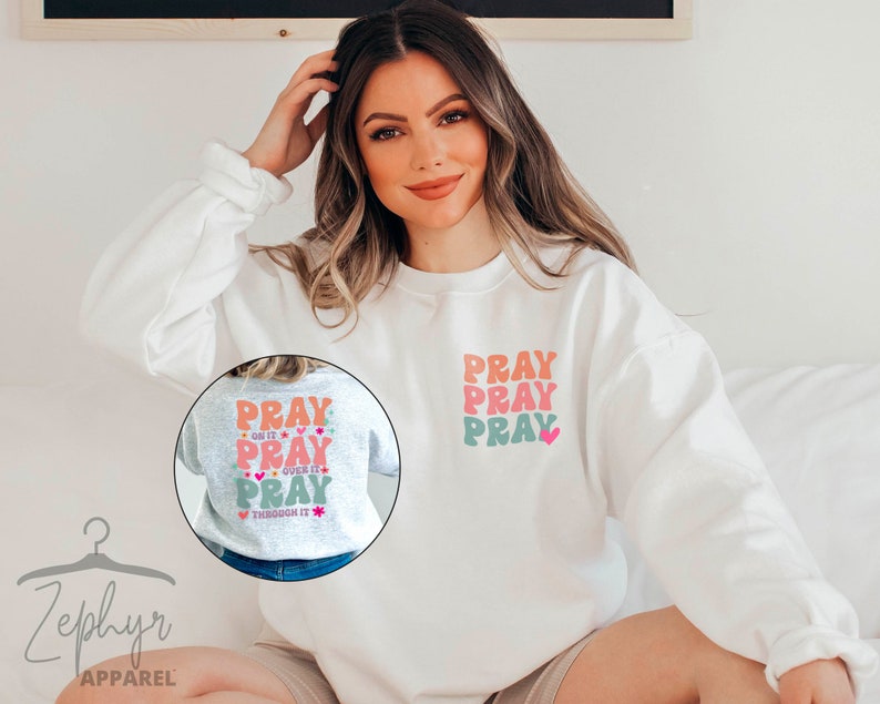 May include: White sweatshirt with a pink, orange and green graphic that says "Pray" three times. The graphic is on the front of the sweatshirt. The sweatshirt is being worn by a person.