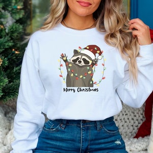 May include: White sweatshirt with a cartoon raccoon wearing a Santa hat and tangled in Christmas lights. The text "Merry Christmas" is printed below the raccoon.