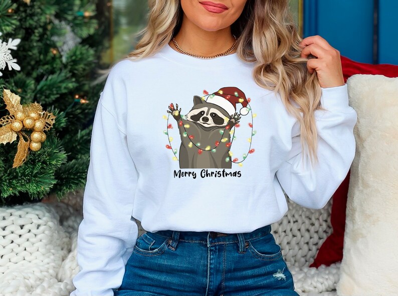 May include: White sweatshirt with a cartoon raccoon wearing a Santa hat and tangled in Christmas lights. The text "Merry Christmas" is printed below the raccoon.