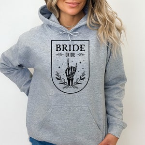 May include: Gray hoodie with a black graphic of a skeleton hand making a rock and roll sign. The graphic is inside a shield with the text "Bride or Die" above the hand.