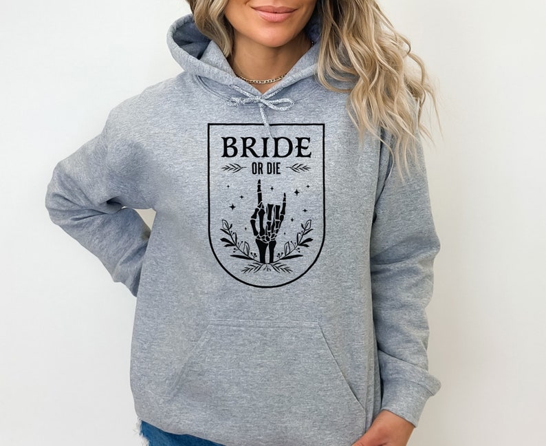May include: Gray hoodie with a black graphic of a skeleton hand making a rock and roll sign. The graphic is inside a shield with the text "Bride or Die" above the hand.