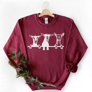 May include: A maroon sweatshirt with a white graphic of three goats peeking over a fence. The goats are looking at the viewer.