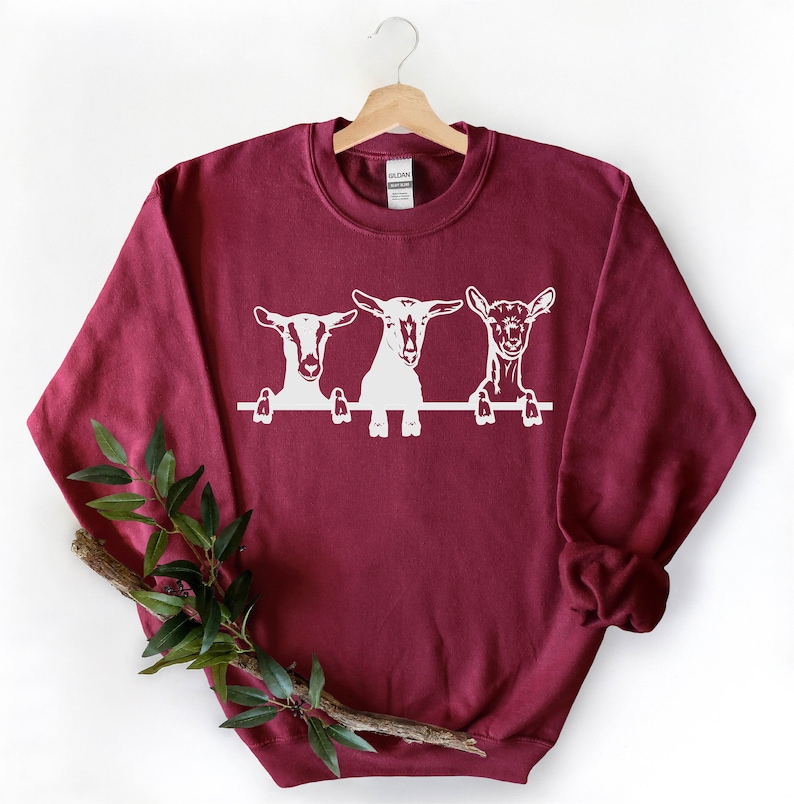 May include: A maroon sweatshirt with a white graphic of three goats peeking over a fence. The goats are looking at the viewer.
