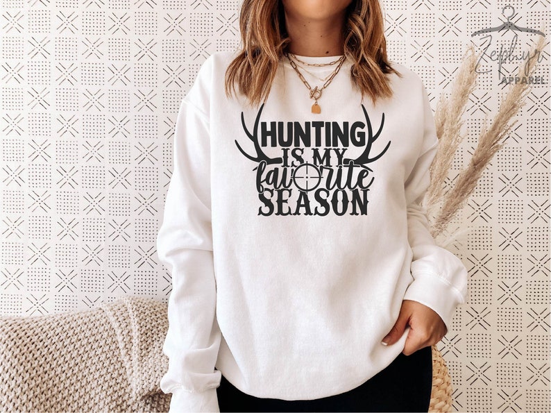 K&ouml;nnte beinhalten: Wei&szlig;er Pullover mit schwarzem Text, der "Hunting is my favorite season" lautet, mit einem Geweih und einem Zielsymbol.