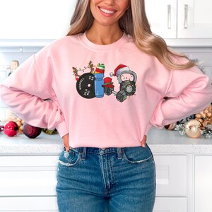 K&ouml;nnte beinhalten: Rosafarbener Pullover mit einem weihnachtlichen Design, das eine Langhantel, eine Kettlebell, eine Hantel und eine Wasserflasche zeigt. Das Design enth&auml;lt Weihnachtslichter, eine Weihnachtsm&uuml;tze und ein Rentier.