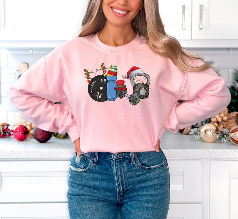K&ouml;nnte beinhalten: Rosafarbener Pullover mit einem weihnachtlichen Design, das eine Langhantel, eine Kettlebell, eine Hantel und eine Wasserflasche zeigt. Das Design enth&auml;lt Weihnachtslichter, eine Weihnachtsm&uuml;tze und ein Rentier.