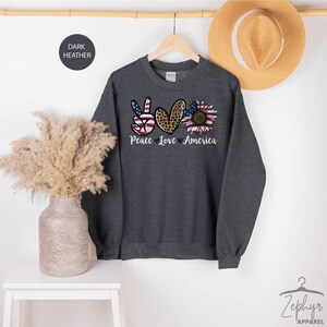May include: Dark heather grey sweatshirt with a peace sign, heart, and sunflower graphic. The peace sign is red, white, and blue, the heart is leopard print, and the sunflower is yellow with red, white, and blue petals. The text "Peace Love America" is printed below the graphic.