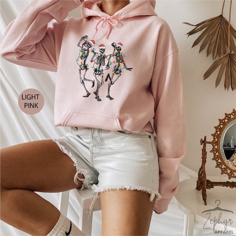 May include: A light pink hoodie with a graphic of three skeletons wearing Santa hats and holding Christmas lights. The hoodie has a drawstring hood and a kangaroo pocket.