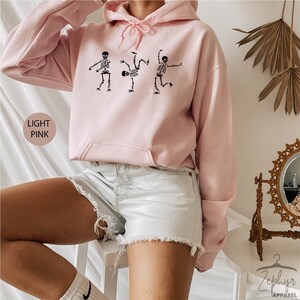 May include: A light pink hoodie with a graphic of three skeletons dancing. The hoodie has a drawstring hood and a kangaroo pocket.