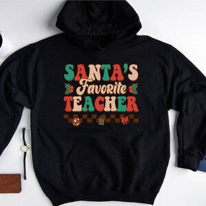 K&ouml;nnte beinhalten: Schwarzer Kapuzenpullover mit einem Retro-Grafik, auf dem "SANTA'S FAVORITE TEACHER" in roter, gr&uuml;ner und wei&szlig;er Schrift steht. Die Grafik enth&auml;lt auch Stechpalmenbl&auml;tter und ein kariertes Muster mit einem Weihnachtsornament, einer Geschenkbox und einer Schleife.