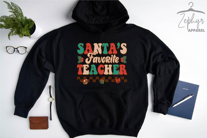 K&ouml;nnte beinhalten: Schwarzer Kapuzenpullover mit einem Retro-Grafik, auf dem "SANTA'S FAVORITE TEACHER" in roter, gr&uuml;ner und wei&szlig;er Schrift steht. Die Grafik enth&auml;lt auch Stechpalmenbl&auml;tter und ein kariertes Muster mit einem Weihnachtsornament, einer Geschenkbox und einer Schleife.