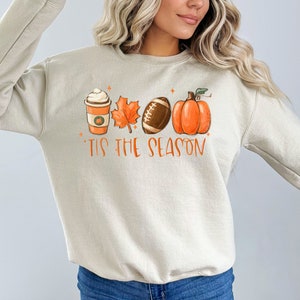 May include: A beige sweatshirt with a graphic design of a coffee cup, a leaf, a football, and a pumpkin. The text "'Tis the season" is printed below the graphic.