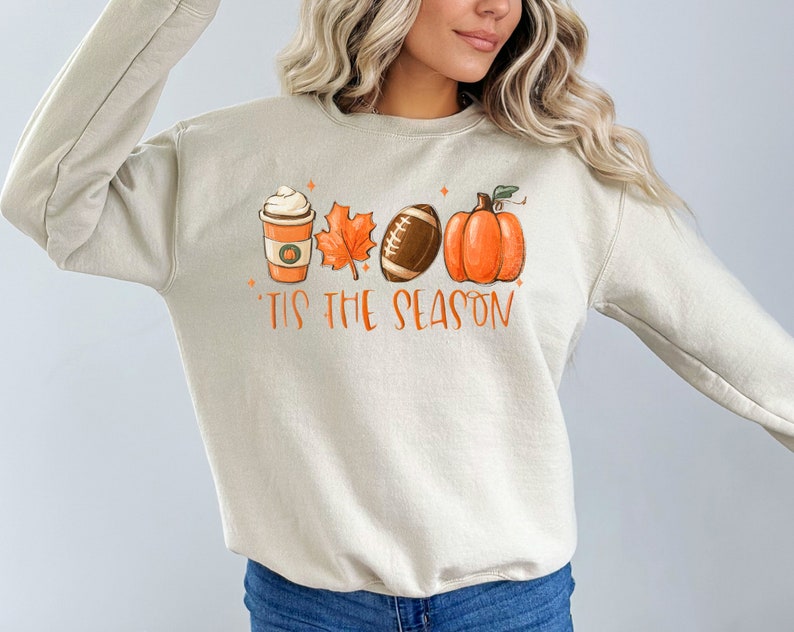 May include: A beige sweatshirt with a graphic design of a coffee cup, a leaf, a football, and a pumpkin. The text "'Tis the season" is printed below the graphic.