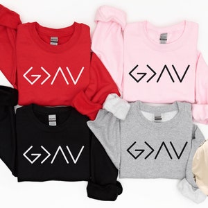 以下が含まれることがあります： 異なる色の4つのスウェットシャツに、前面に「G>V」という文字が印刷されています。ベージュのスウェットシャツには、背面に「神は高みと低みよりも偉大である」という文字が印刷されています。
