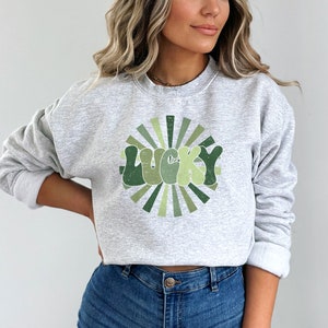 Peut inclure: Un sweat-shirt gris avec un motif vert et blanc du mot "LUCKY" dans une police r&eacute;tro avec un design en forme de soleil.