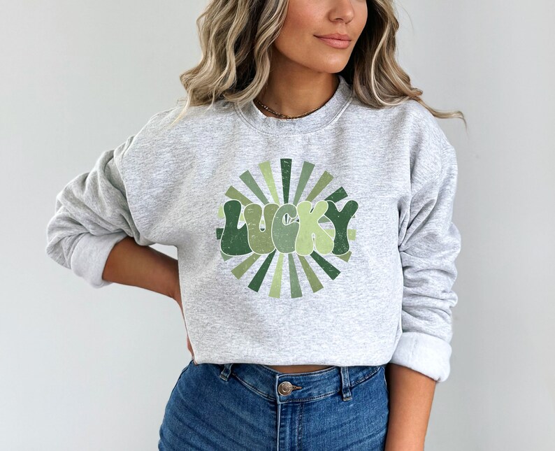 Peut inclure: Un sweat-shirt gris avec un motif vert et blanc du mot "LUCKY" dans une police r&eacute;tro avec un design en forme de soleil.