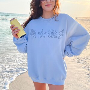 May include: A light blue sweatshirt featuring a line of seashell illustrations. The sweatshirt is worn on a beach with the ocean in the background. A yellow insulated water bottle is held in the hand.