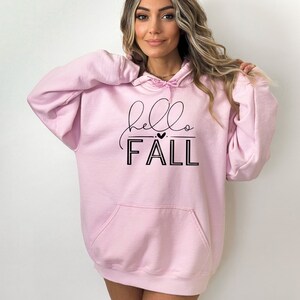 Op de afbeelding: Roze oversized hoodie met een zwarte grafische afbeelding die "hello FALL" zegt met een klein hartje boven het woord "FALL".