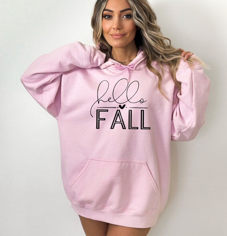 Op de afbeelding: Roze oversized hoodie met een zwarte grafische afbeelding die "hello FALL" zegt met een klein hartje boven het woord "FALL".