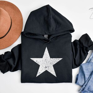 May include: Black hooded sweatshirt with a white distressed star graphic. The sweatshirt is laid flat on a white surface with a brown hat and blue jeans.