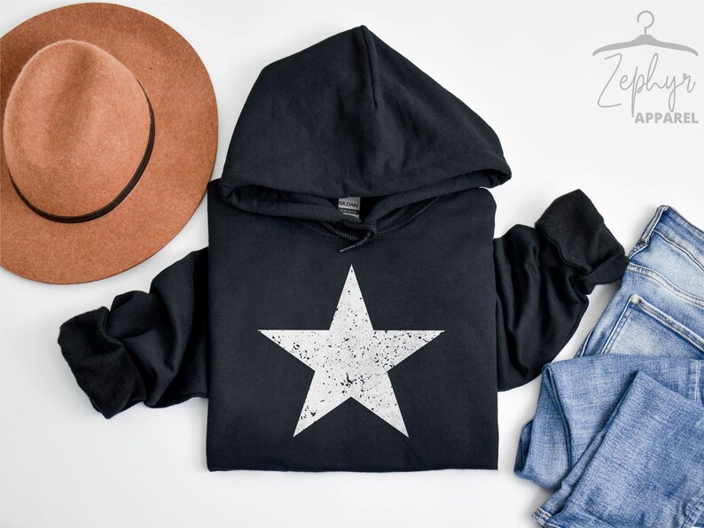 May include: Black hooded sweatshirt with a white distressed star graphic. The sweatshirt is laid flat on a white surface with a brown hat and blue jeans.
