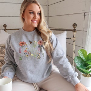 May include: A gray sweatshirt with a floral print featuring butterflies and leaves. The sweatshirt is a crew neck style with long sleeves. The text "SPORTS GREY" is visible in the lower right corner of the image.