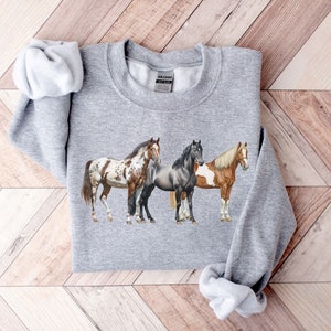 May include: A gray sweatshirt with a print of three horses. The horses are a brown and white spotted horse, a gray horse, and a brown and white horse. The horses are standing side by side.