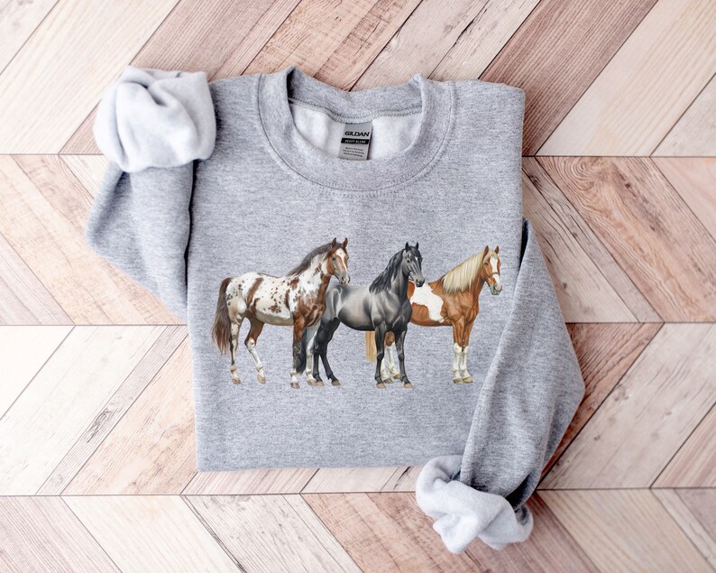 May include: A gray sweatshirt with a print of three horses. The horses are a brown and white spotted horse, a gray horse, and a brown and white horse. The horses are standing side by side.