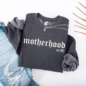 Op de afbeelding: Een donkergrijs sweatshirt met de tekst "motherhood est. 2025" in witte gotische letters. Het sweatshirt is gecombineerd met lichtblauwe jeans.
