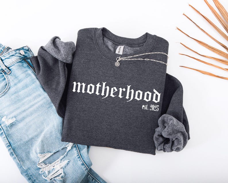 Op de afbeelding: Een donkergrijs sweatshirt met de tekst "motherhood est. 2025" in witte gotische letters. Het sweatshirt is gecombineerd met lichtblauwe jeans.